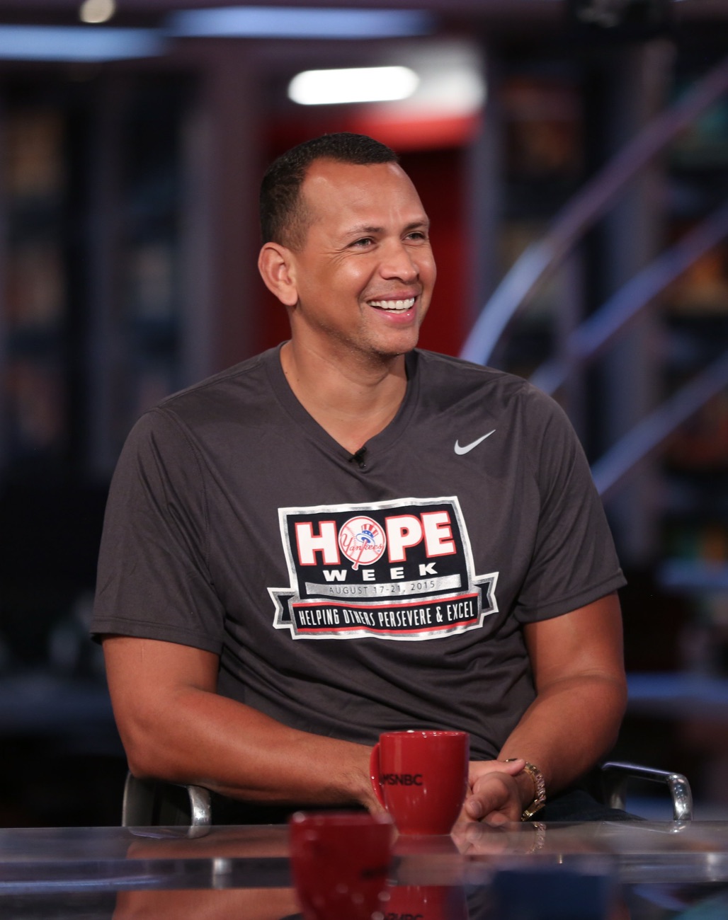 Alex Rodriguez laughs at a studio desk in a gray Hope Week t-shirt.