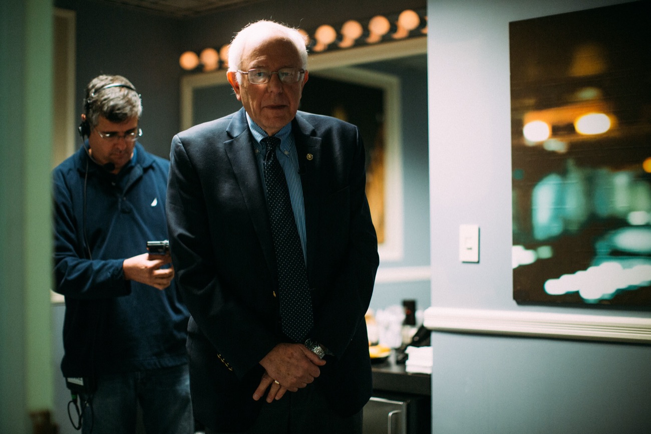 Bernie Sanders waits in a backstage dressing room beside a staffer checking a phone.