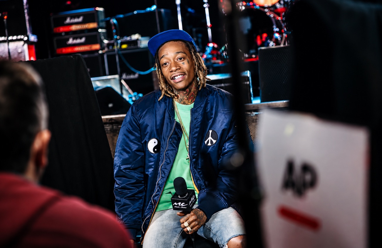 Wiz Khalifa, in a blue jacket and cap, sits on stage holding a microphone.