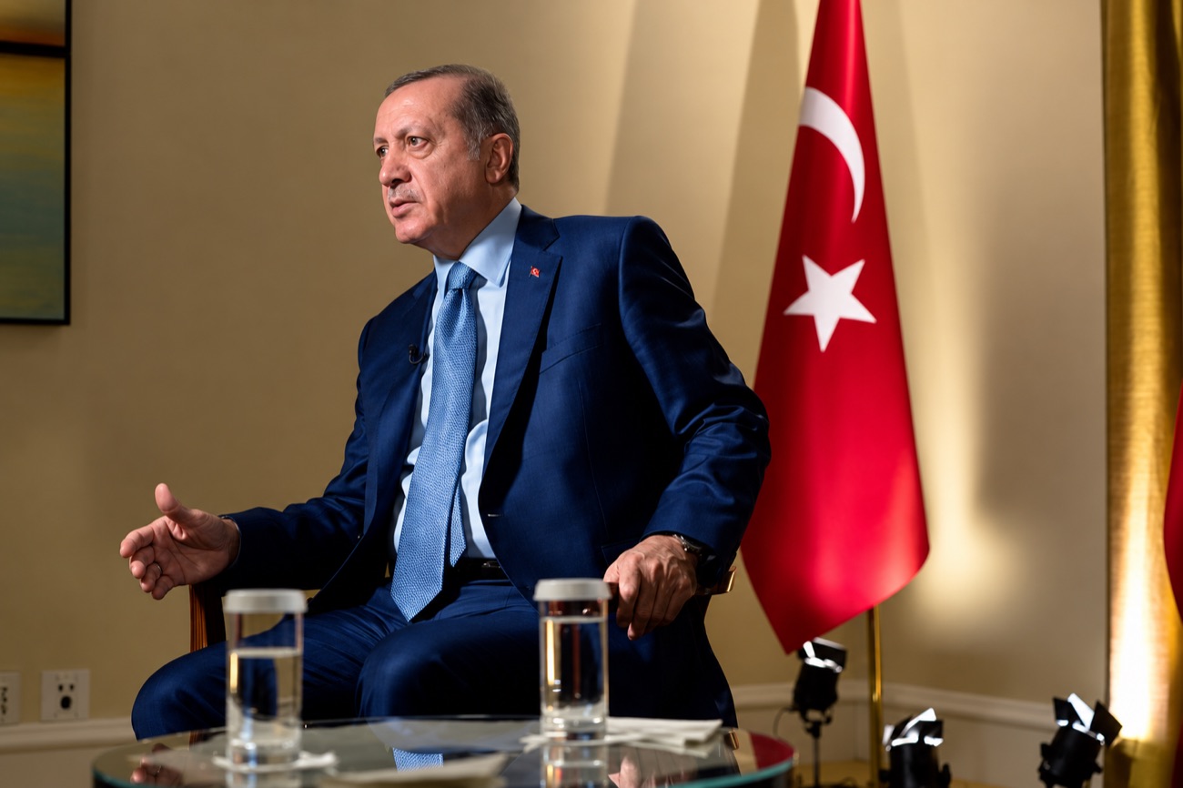 Turkish President Recep Tayyip Erdoğan gestures during a seated interview beside the Turkish flag.