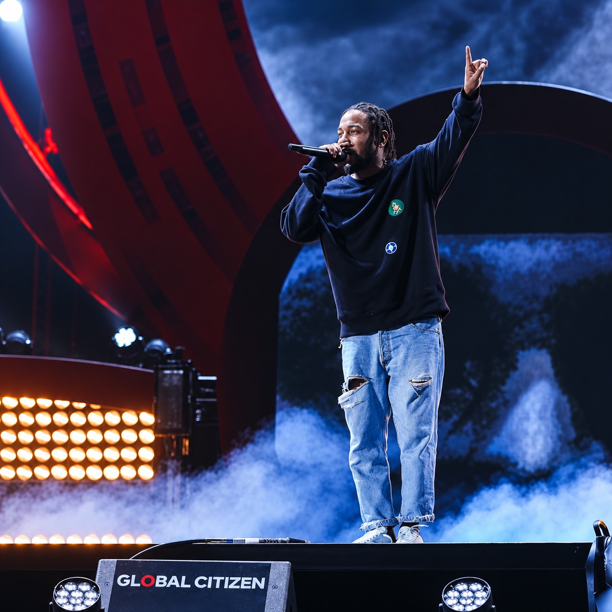 Kendrick Lamar raises one arm on a smoky Global Citizen stage.