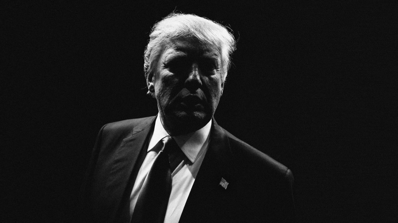 A low-key black and white portrait of President Donald Trump in a dark suit, his face half in shadow.