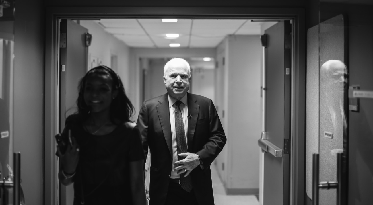 John McCain walks out of a narrow studio corridor, in black and white.