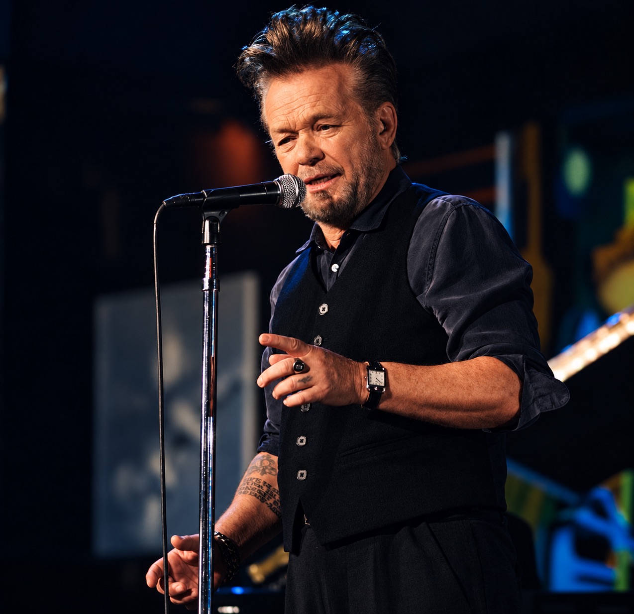 John Mellencamp sings into a stand microphone on a dark stage.