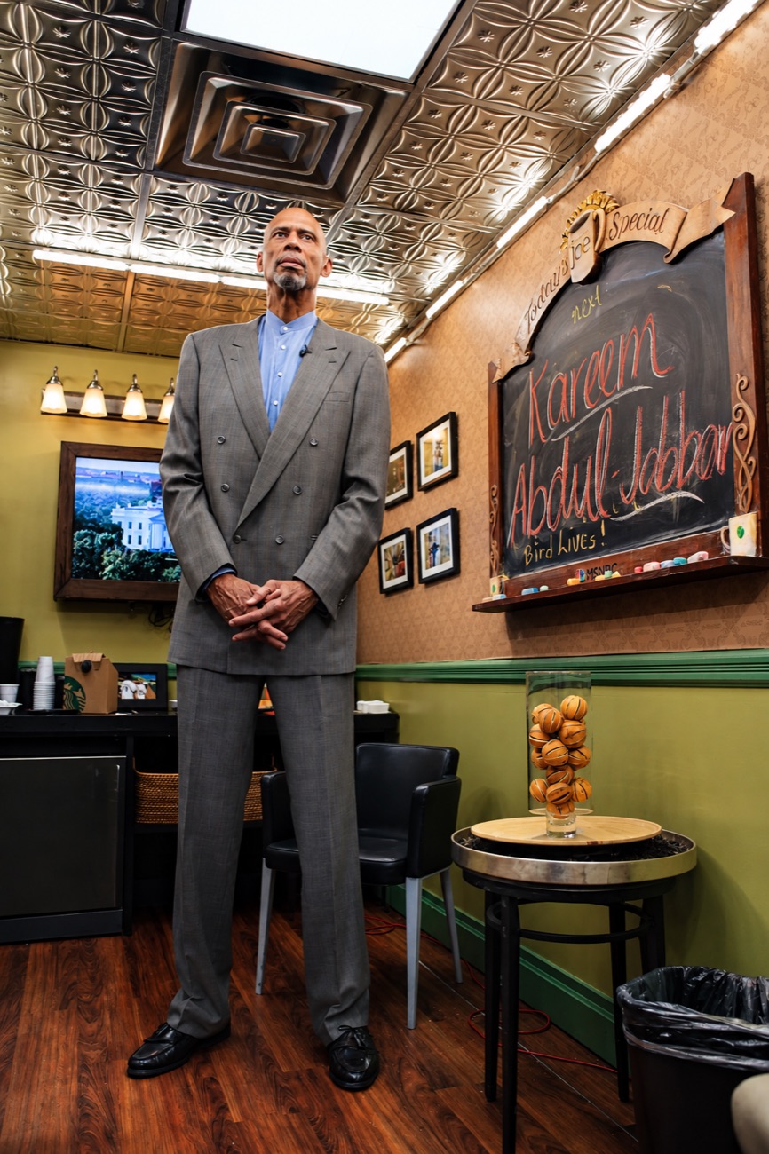 Kareem Abdul-Jabbar stands in a gray suit in a wood-paneled room, his name chalked on a board behind him.