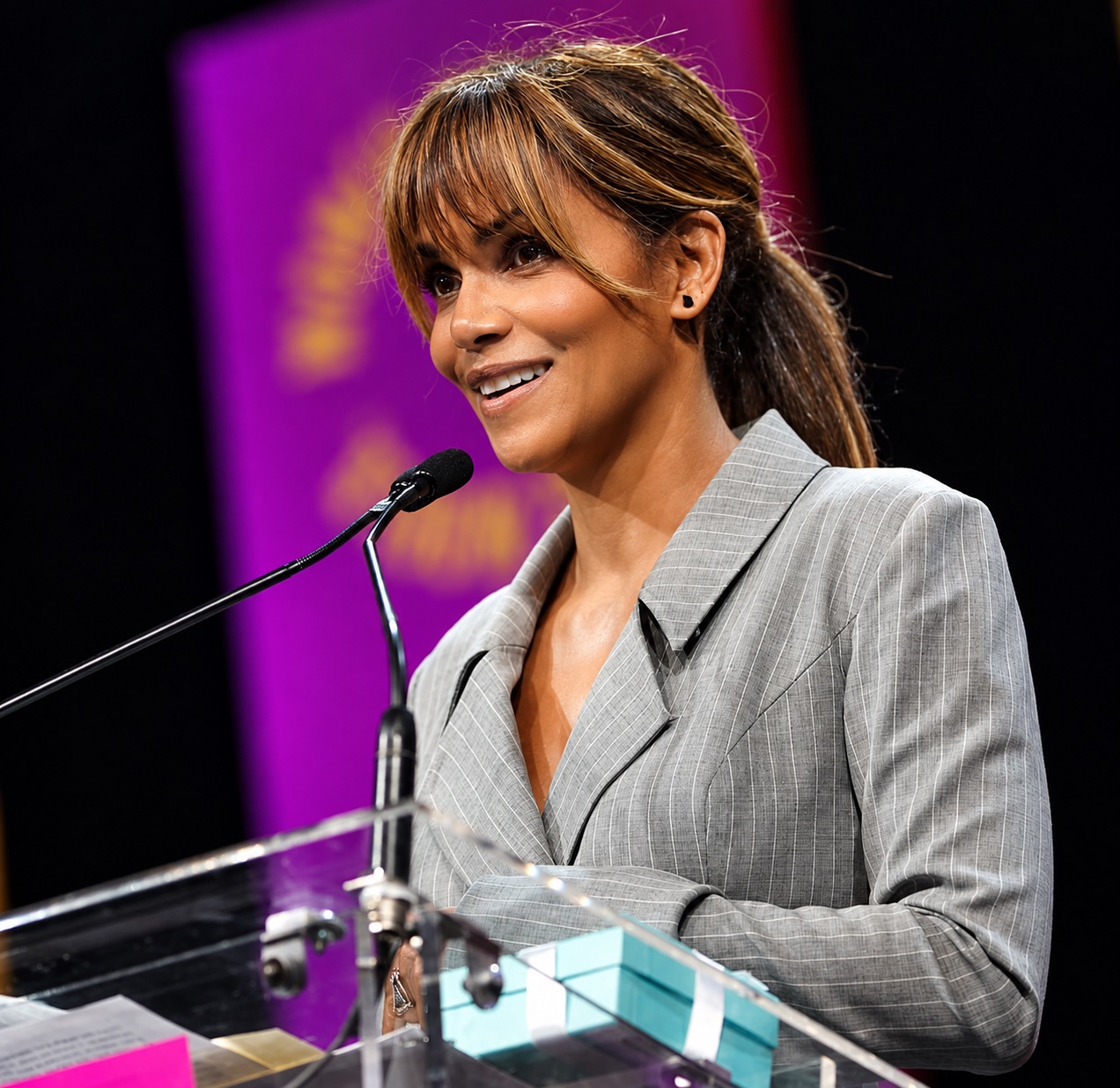 Halle Berry, in a grey pinstripe blazer, speaks at a podium microphone.