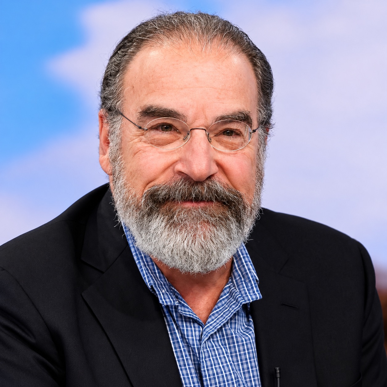 Mandy Patinkin smiles in a dark jacket and checked shirt against a soft blue backdrop.