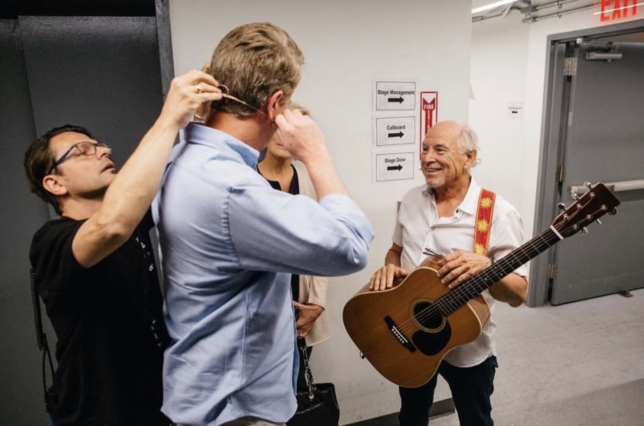 Jimmy Buffett, guitar in hand, smiles backstage as a guest is fitted with a microphone.