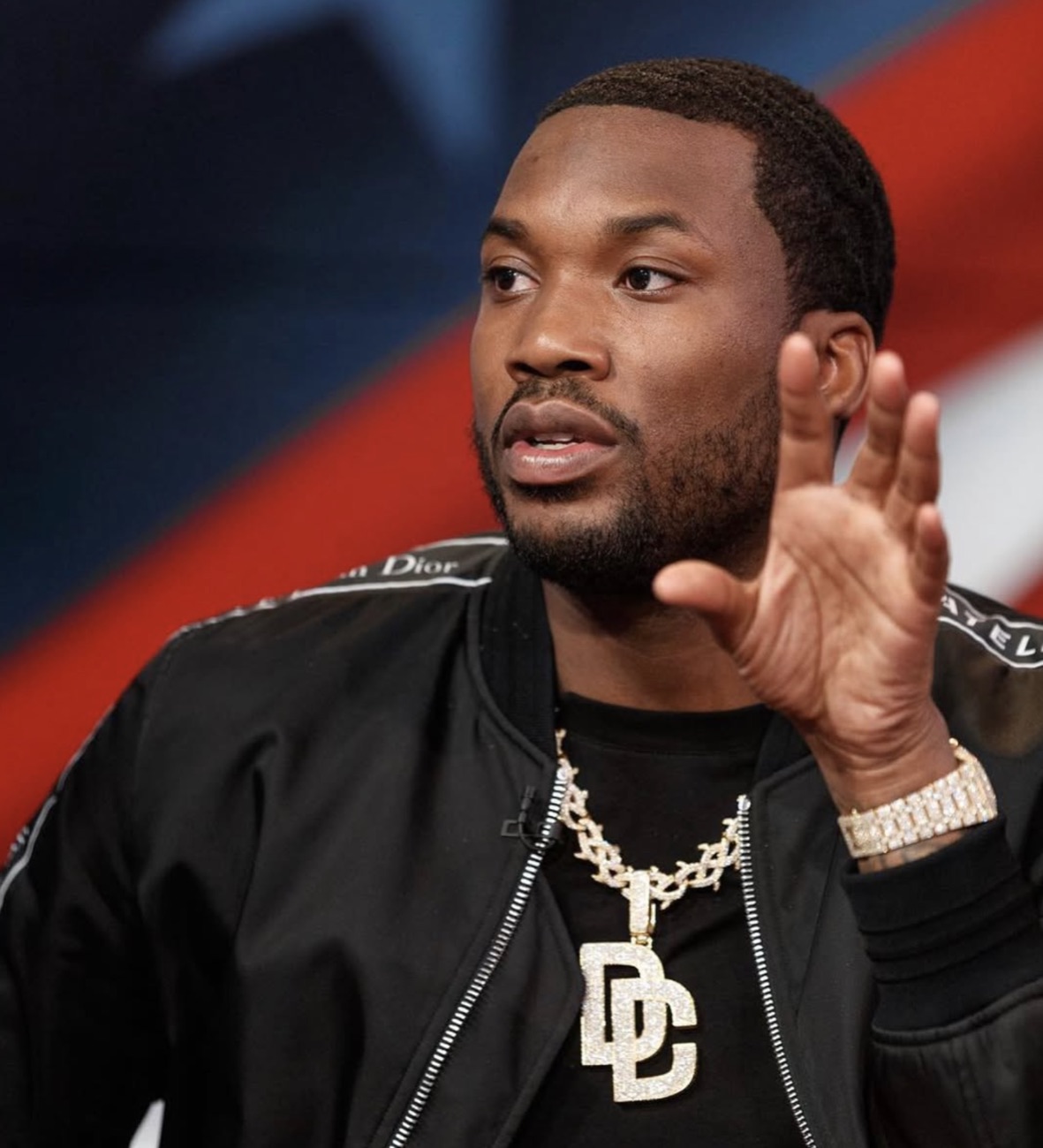 Meek Mill, in a black jacket and gold chain, gestures mid-sentence in a studio.