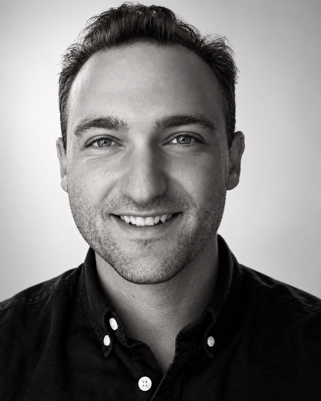 Miller Hawkins smiling in a black button-down shirt, in a black-and-white studio portrait.