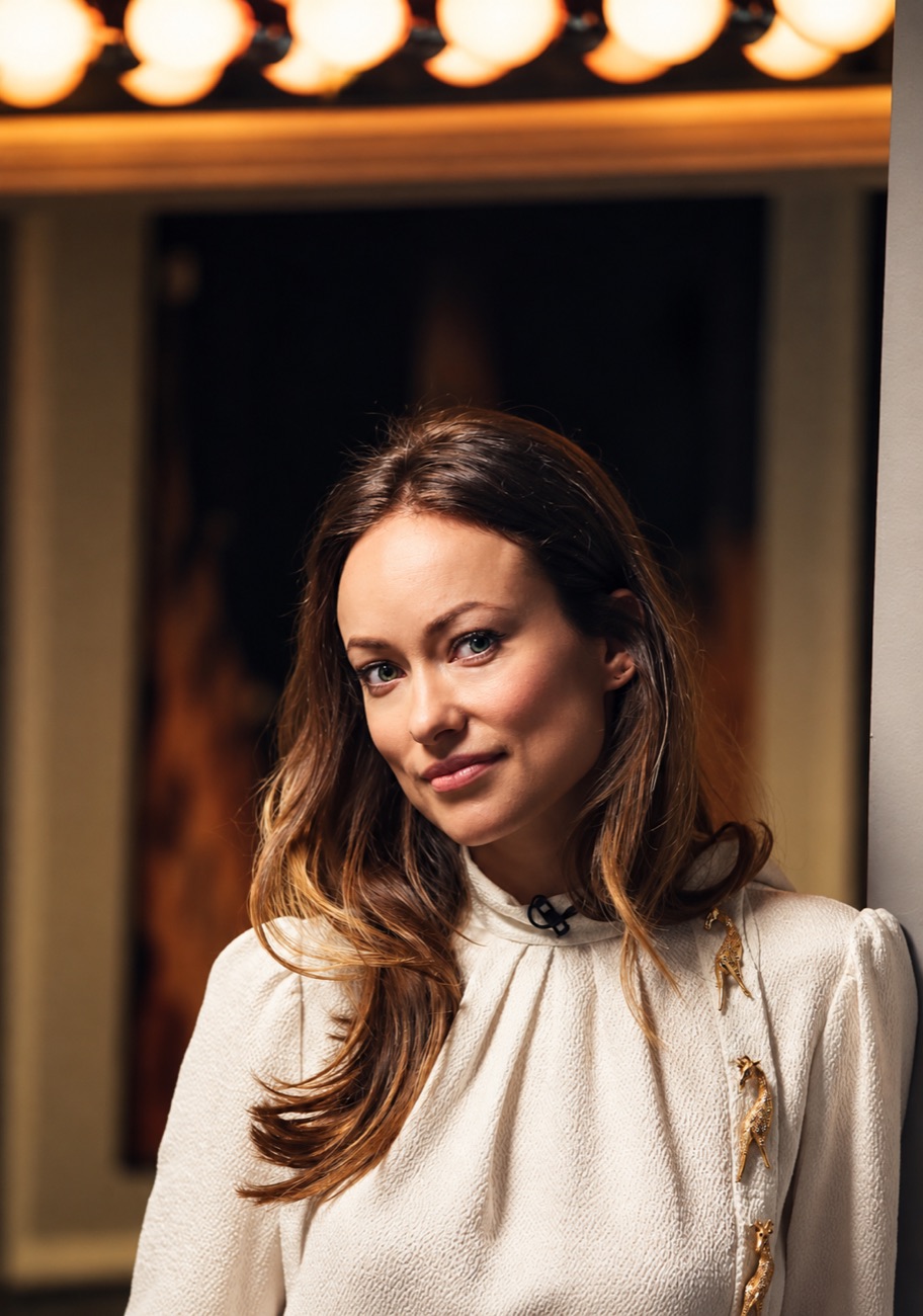 Olivia Wilde leans against a mirror lined with vanity bulbs, in a cream blouse.
