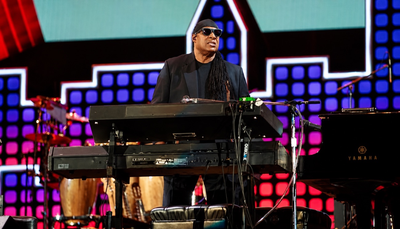 Stevie Wonder plays a bank of keyboards on a stage lit red, white and blue.