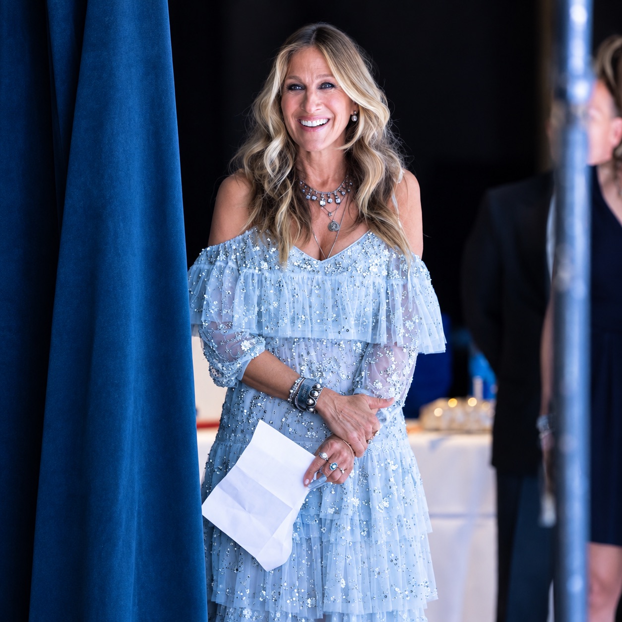 Sarah Jessica Parker, in a beaded white dress, waits beside a blue-lit stage curtain with cue cards in hand.