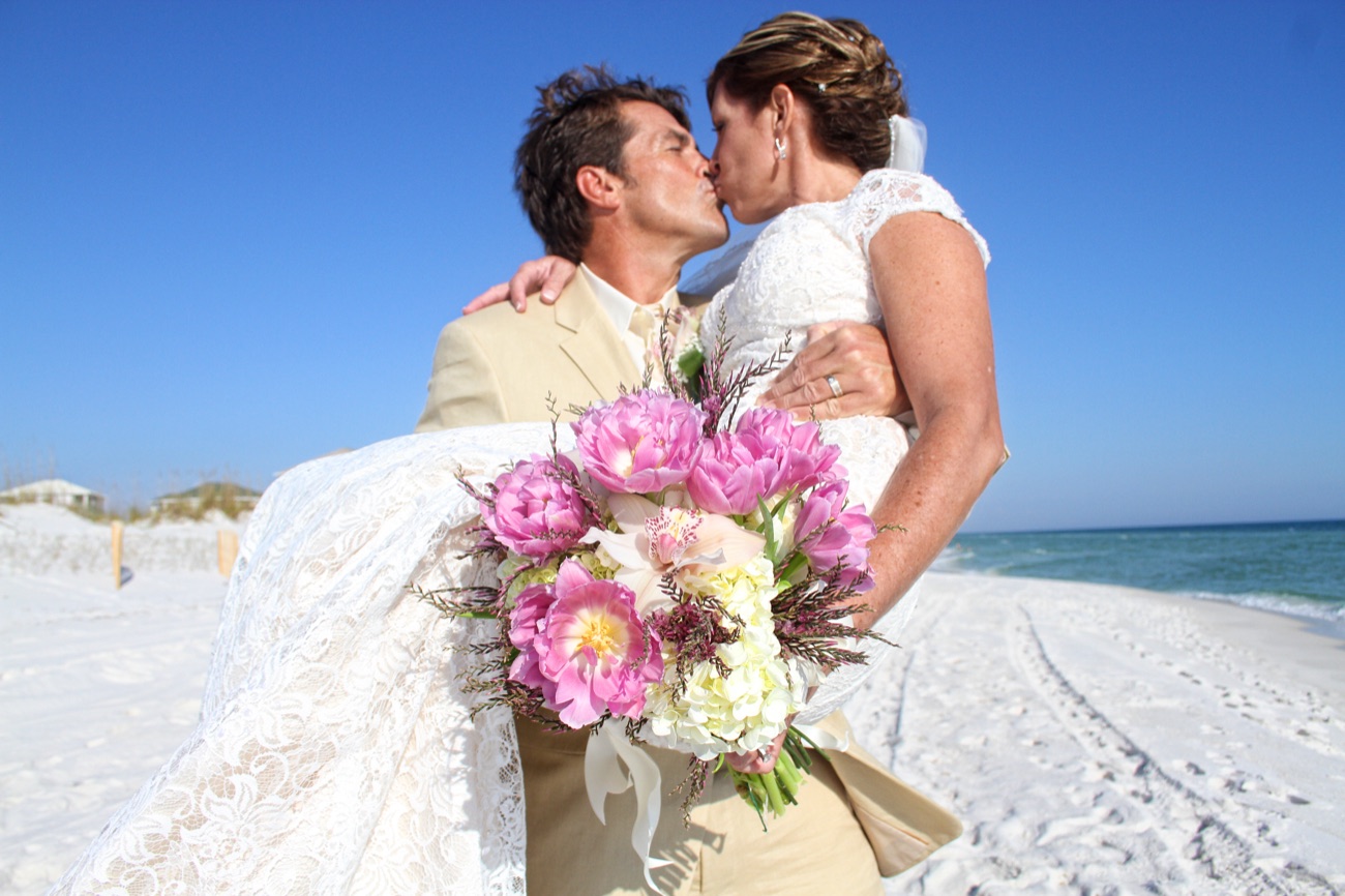 A groom carries his bride across white sand and kisses her, a bouquet of pink peonies in her hand.