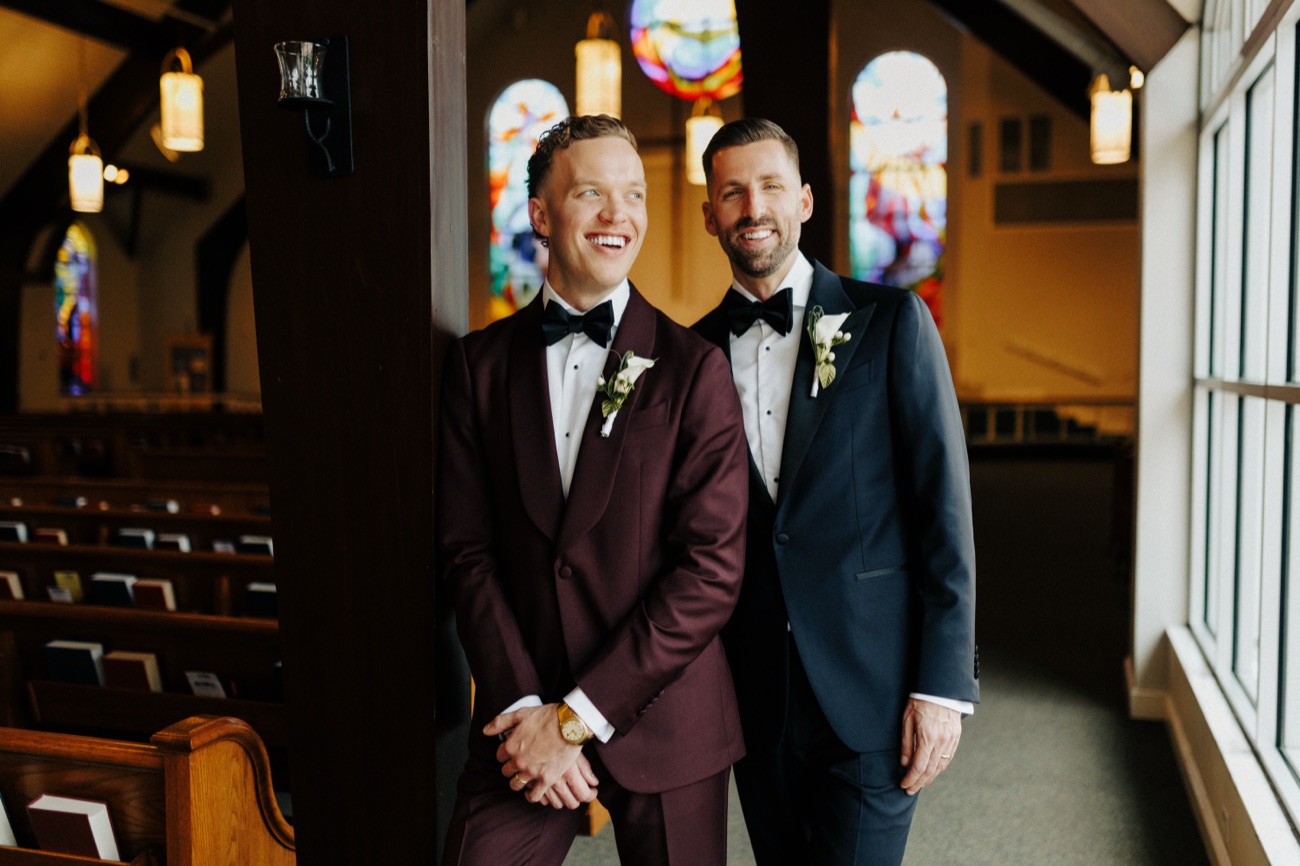 Two grooms in tuxedos, one in burgundy velvet, laugh together in a sunlit chapel.