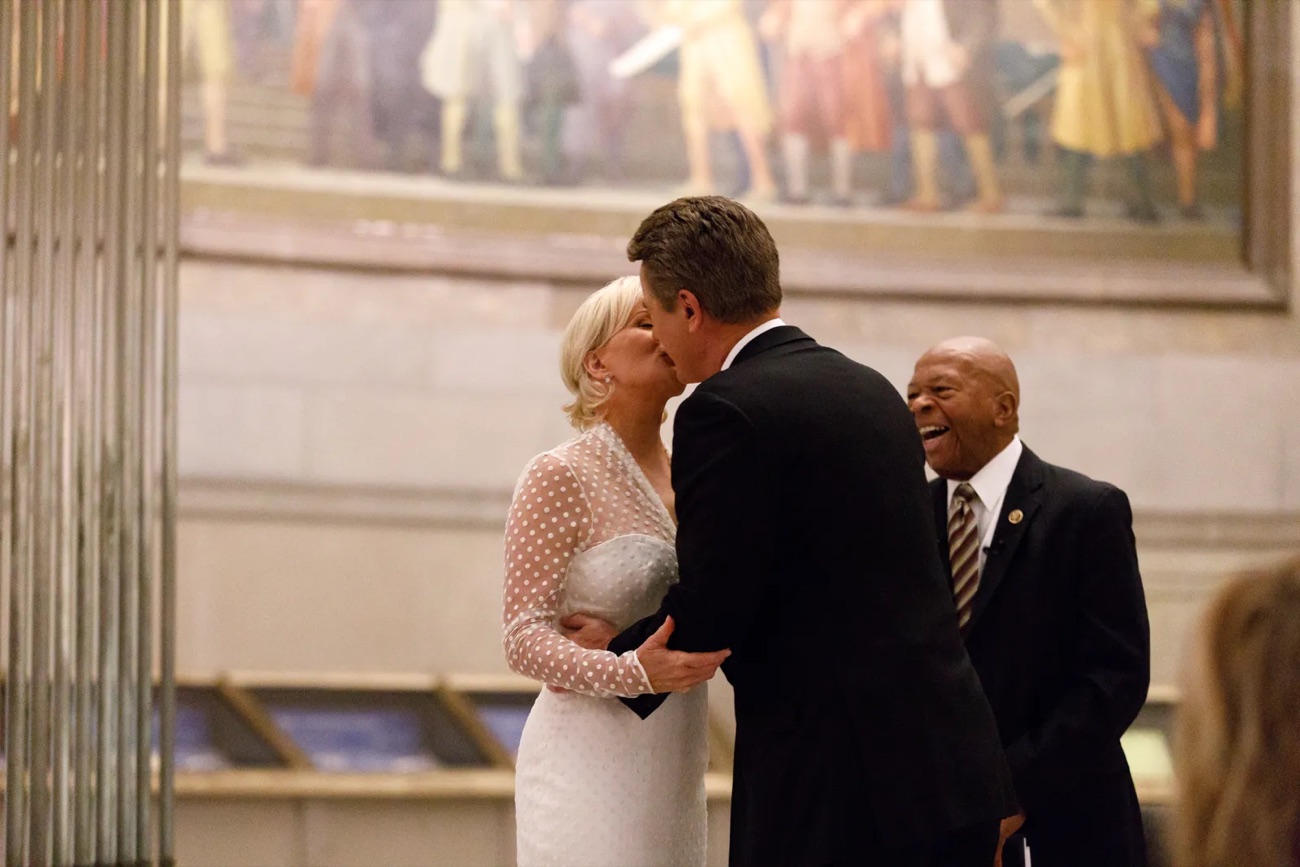 A groom kisses his bride as the officiant laughs beneath a historic mural.