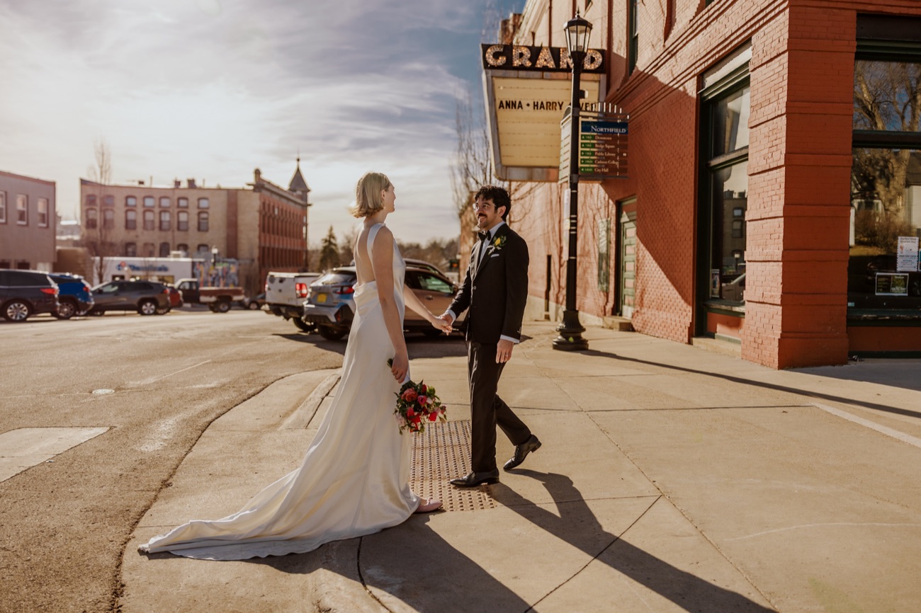 A bride and groom hold hands on a sunny street corner beneath a theater marquee bearing their names.