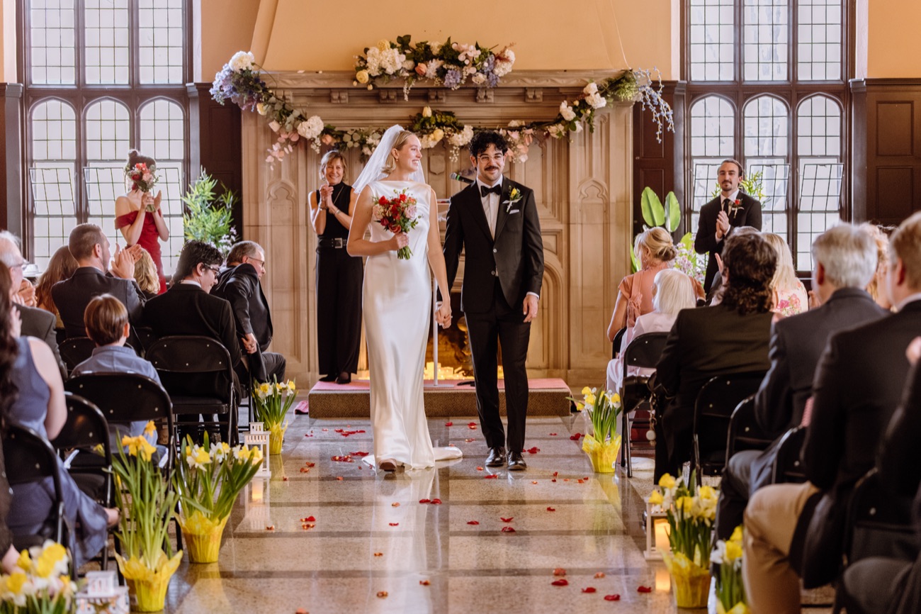 A bride and groom walk back up the aisle hand in hand through a grand hall of applauding guests, daffodils and rose petals.