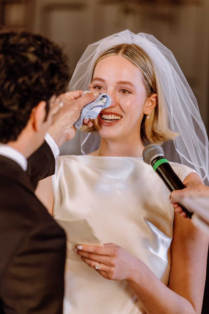 A groom wipes happy tears from his bride's face with a handkerchief as she reads her vows.
